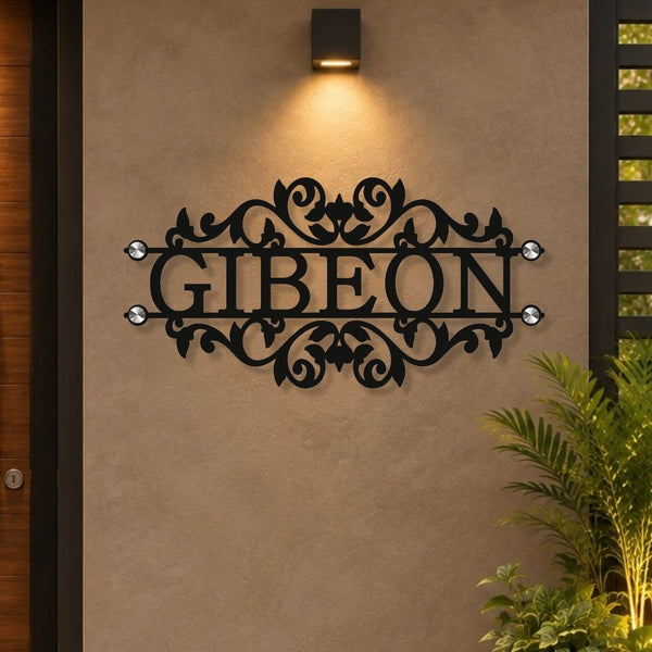 Black metal personalized name sign reading 'GIBEON' with ornate scrollwork and small clear stone accents, mounted on a beige stucco wall under a wall light.