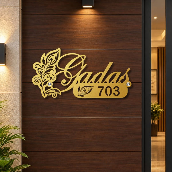 Gold metal house number sign reading 'Gadas 703' with decorative leaf flourishes, mounted on a dark wood-paneled exterior wall.