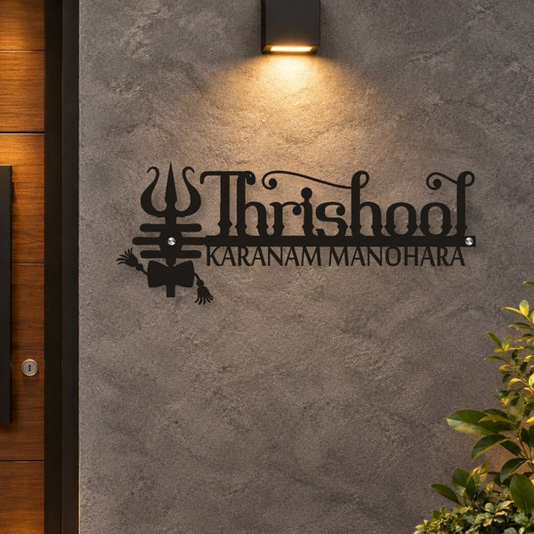 Black metal wall art featuring the word 'Thrisool' and the text 'KARANAM MANOHARA' with a trident motif, mounted on a textured gray concrete wall under a lit sconce.
