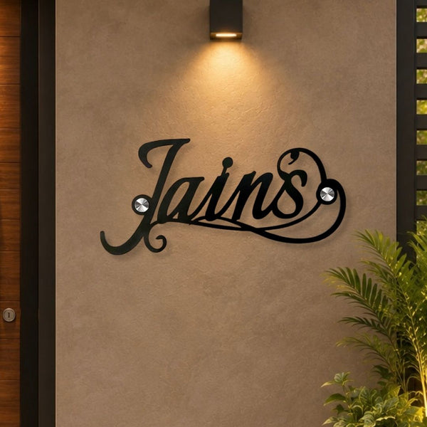 Black metal script wall sign spelling 'Jains' mounted on a beige stucco wall, with a warm wall sconce above and green plants at the lower right.