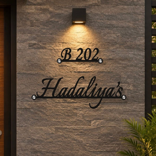 Black metal personalized house number and name sign mounted on a gray stone wall, with a warm wall light above and green foliage in the lower right corner.