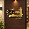 Gold-toned metal wall art featuring Devanagari script, a seated figure silhouette, and the names 'VARNIKA & TARUN' with address 'T4 35 A', mounted on a dark wood-paneled wall in an interior setting.
