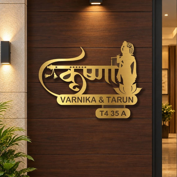 Gold-toned metal wall art featuring Devanagari script, a seated figure silhouette, and the names 'VARNIKA & TARUN' with address 'T4 35 A', mounted on a dark wood-paneled wall in an interior setting.
