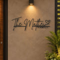 Personalized black metal wall art reading 'The Matthew Family' with a heart, mounted on a textured beige stucco wall under a warm wall sconce, with a plant in the foreground.