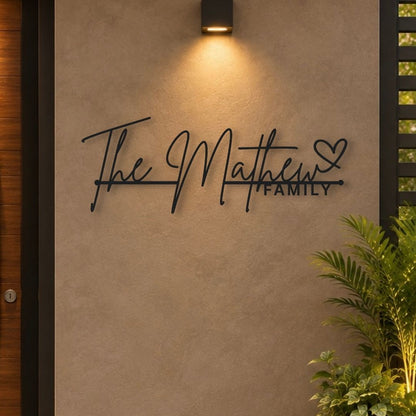 Personalized black metal wall art reading 'The Matthew Family' with a heart, mounted on a textured beige stucco wall under a warm wall sconce, with a plant in the foreground.