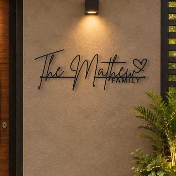 Personalized black metal wall art reading 'The Matthew Family' with a heart, mounted on a textured beige stucco wall under a warm wall sconce, with a plant in the foreground.