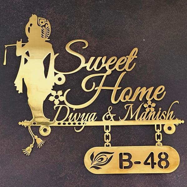 Gold-colored laser-cut metal sign featuring a silhouette of Radha Krishna, the script text 'Sweet Home Divya & Manish', and a hanging plaque with 'B-48', displayed against a dark textured wall.
