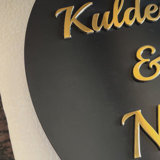 Modern metal house number and name sign mounted on a beige wall, featuring a black circular nameplate with gold lettering and a separate vertical silver number plate next to it.

