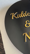 Modern metal house number and name sign mounted on a beige wall, featuring a black circular nameplate with gold lettering and a separate vertical silver number plate next to it.

