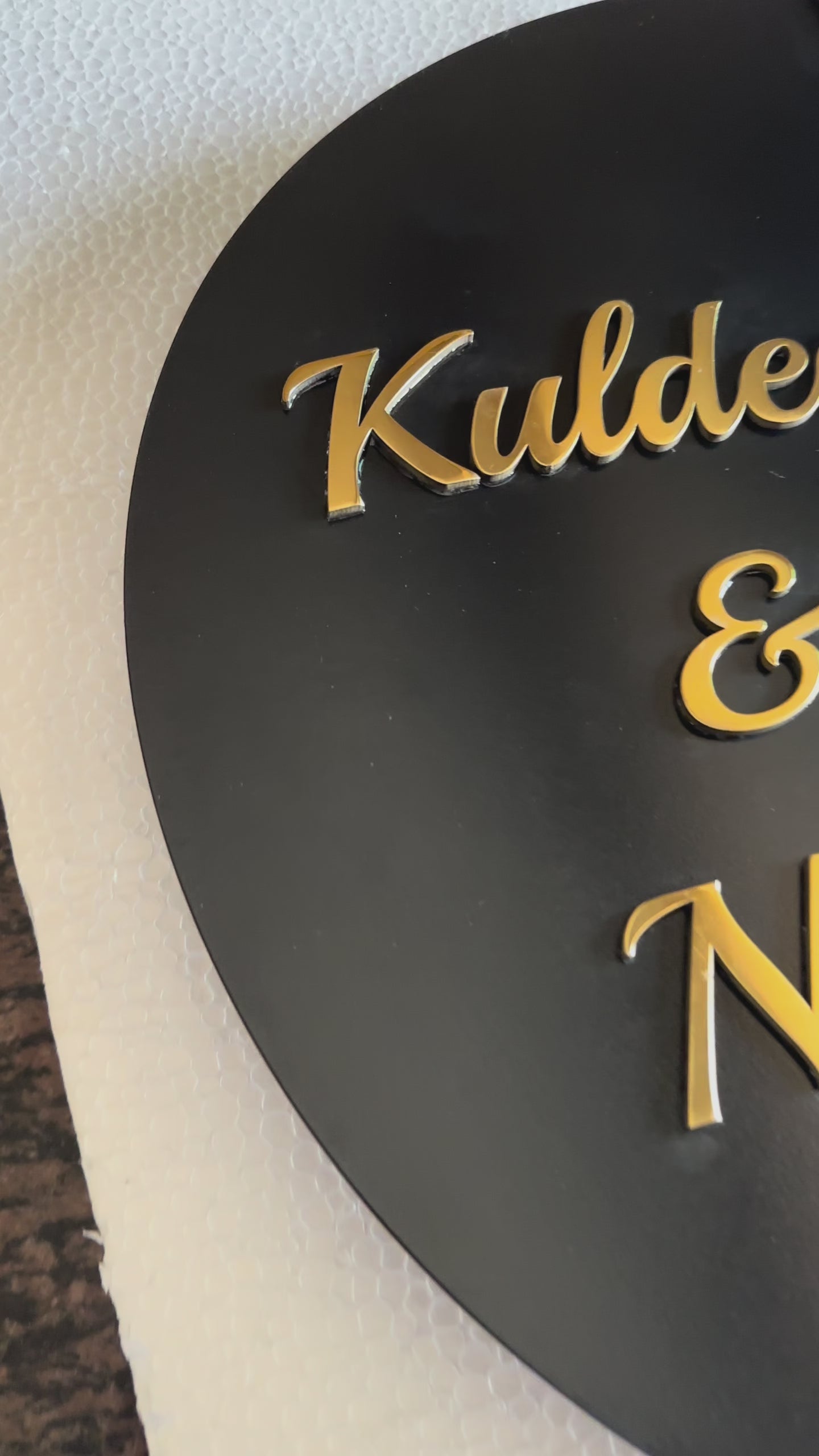 Modern metal house number and name sign mounted on a beige wall, featuring a black circular nameplate with gold lettering and a separate vertical silver number plate next to it.

